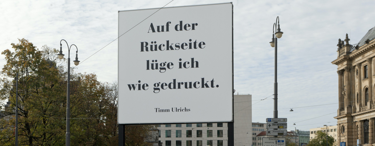 Schräge Ansicht des Billboards. In großen schwarzen Buchstaben auf weißem Grund zu lesen: "Auf der Rückseite lüge ich wie gedruckt. Timm Ulrichs".